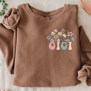 以下が含まれることがあります： 花柄のデザインとカラフルな文字で「GIGI」と書かれた茶色のスウェットシャツです。