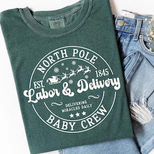 May include: A blue spruce t-shirt with a white circular graphic that reads "North Pole Labor & Delivery Baby Crew" with reindeer and a sleigh. The shirt is paired with denim jeans and a small bouquet of white flowers.