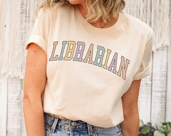 May include: A woman wearing a light beige t-shirt with the text "LIBRARIAN" in rainbow colors. She is wearing blue jeans with distressed details.