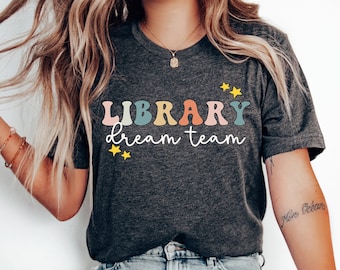 May include: Dark grey heather t-shirt with the words "LIBRARY dream team" in colorful, retro-style lettering. The design includes yellow star accents. The shirt is a crew neck and has short sleeves.