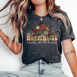 May include: A dark grey heather t-shirt with a floral design and the text "NORMALIZE minds of all kinds".