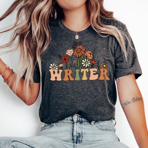 May include: Dark grey heather t-shirt with a floral design and the word "WRITER" in a retro font.