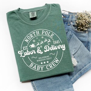 May include: A light green t-shirt with the text "NORTH POLE Labor & Delivery BABY CREW" in a circular design. The design includes snowflakes, reindeer, and a sleigh. The shirt is paired with blue denim shorts and a small bouquet of white flowers.