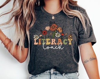 May include: Dark grey heather t-shirt with a floral design and the words "Literacy Coach" in a retro font. The shirt features a bouquet of orange, yellow, and white flowers. The text "Dark Grey Heather" is at the bottom of the image.