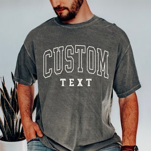 May include: A dark gray t-shirt with a white outline of the word "CUSTOM" in a collegiate font. The word "TEXT" is printed below the word "CUSTOM" in a smaller font.