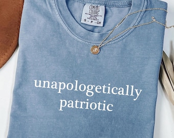 May include: A blue jean colored t-shirt with the words "unapologetically patriotic" in white. A gold necklace with a small pendant rests on the shirt. The shirt is folded and has a tag that says "Comfort Colors".