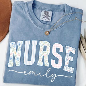 May include: Blue jean colored t-shirt with the word "NURSE" in floral and striped patterns, and the name "emily" in a cursive font. A gold necklace with a pendant rests on the shirt.