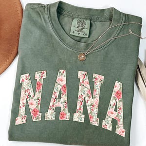 May include: A moss green t-shirt with the word "NANA" in large, floral-patterned letters. A gold necklace with a pendant rests on the shirt. The shirt is folded, and a brown hat is visible in the background.