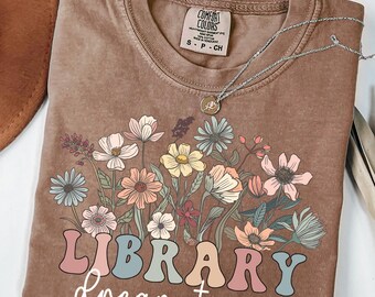 May include: Espresso-colored t-shirt with a floral design and the words "LIBRARY dream team" in a retro font. The shirt features a crew neck and short sleeves. A silver necklace with a pendant is draped across the shirt.