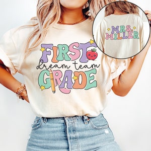 May include: A white t-shirt with the text "First Dream Team Grade" in colorful, retro-style lettering. The shirt also features a cartoon apple and pencil. The text "Mrs. Miller" is visible on the back of the shirt.