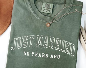 May include: A moss green t-shirt with the text "JUST MARRIED 50 YEARS AGO" in white block letters. A gold-colored necklace with a pendant rests on the shirt. A brown hat and dried plant are in the background.