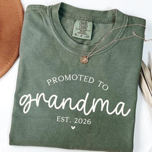 May include: A moss green t-shirt with the words "Promoted to grandma" in white script, along with "Est. 2026" and a small heart. A gold necklace with a pendant rests on the shirt. The shirt is folded.