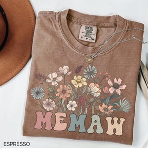May include: A brown t-shirt with a floral design and the text "MEMAW" in colorful letters. The shirt is made of a soft, comfortable material and has a relaxed fit.