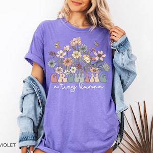 May include: Violet-colored t-shirt with the text "Growing a tiny human" in a retro font. The shirt features a floral design with butterflies. The shirt is made of a soft material.