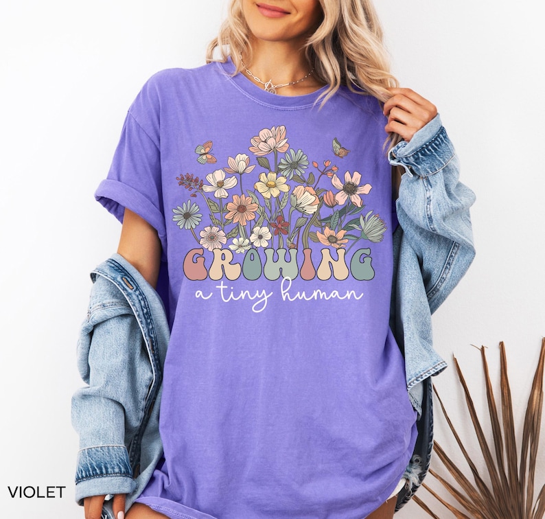May include: Violet-colored t-shirt with the text "Growing a tiny human" in a retro font. The shirt features a floral design with butterflies. The shirt is made of a soft material.