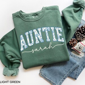 May include: A light green sweatshirt with the word "AUNTIE" in white block letters filled with a blue floral pattern. The name "sarah" is written in cursive below. The sweatshirt is folded on top of a pair of blue jeans.