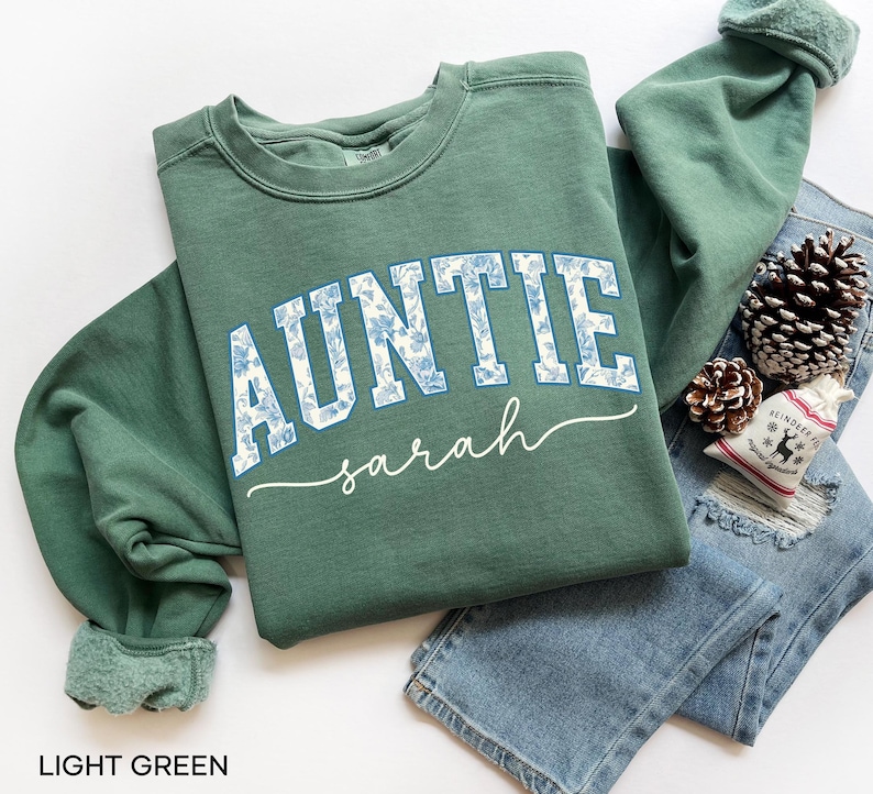 May include: A light green sweatshirt with the word "AUNTIE" in white block letters filled with a blue floral pattern. The name "sarah" is written in cursive below. The sweatshirt is folded on top of a pair of blue jeans.