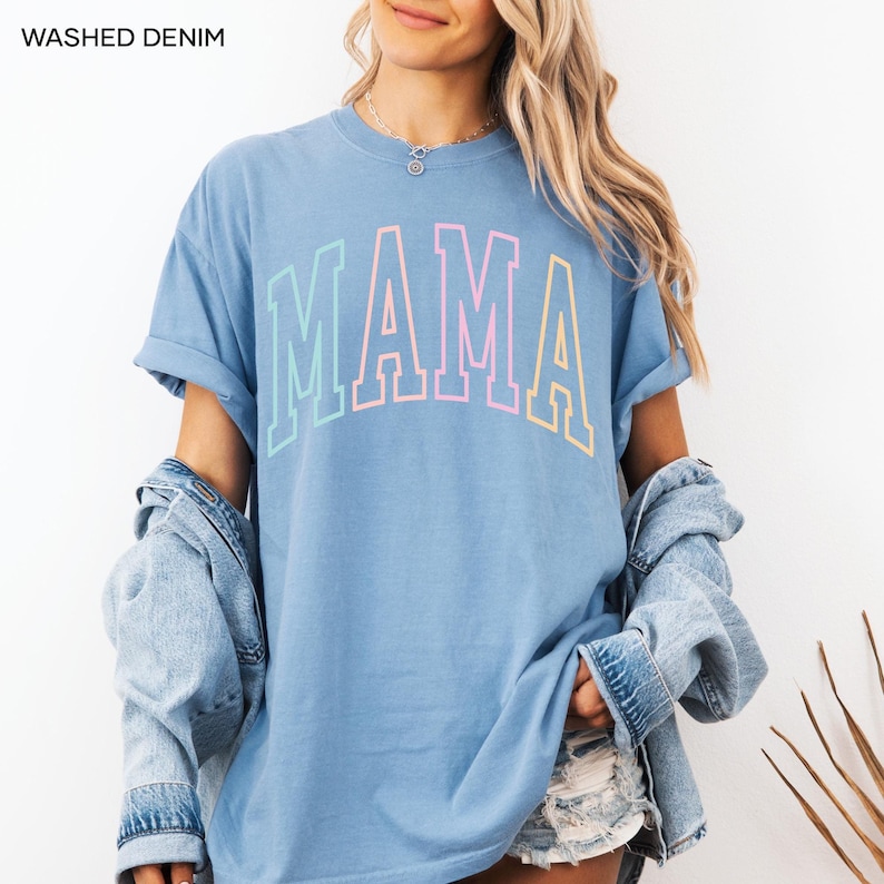 May include: A light blue t-shirt with the word "MAMA" printed in rainbow colors. The shirt has a relaxed fit and rolled up sleeves.