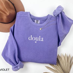 May include: A violet sweatshirt with the word "doula" in white script, a small heart below the word, and a gold necklace. A brown hat is visible in the background. The sweatshirt has long sleeves with rolled cuffs.