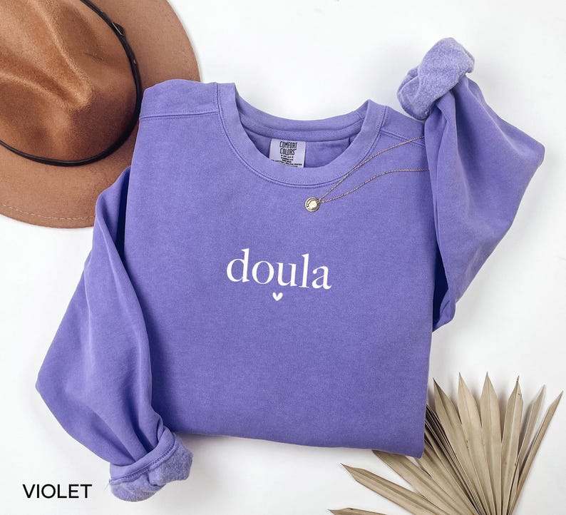 May include: A violet sweatshirt with the word "doula" in white script, a small heart below the word, and a gold necklace. A brown hat is visible in the background. The sweatshirt has long sleeves with rolled cuffs.