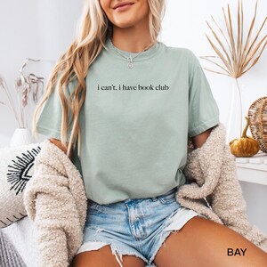 May include: A light green t-shirt with the text "i can't, i have book club" printed on the front.