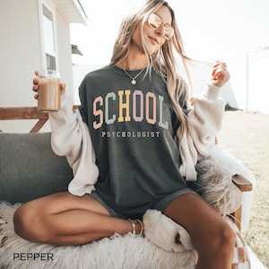 May include: A dark gray sweatshirt with the text "SCHOOL PSYCHOLOGIST" in colorful letters. The sweatshirt is worn by a person with long blonde hair and sunglasses.