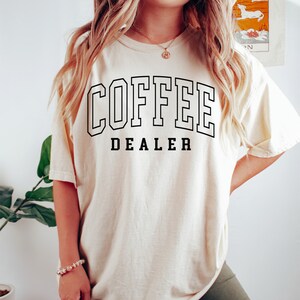May include: Ivory-colored t-shirt with the words "COFFEE DEALER" in black block letters. The text is outlined in black. The shirt is a relaxed fit.