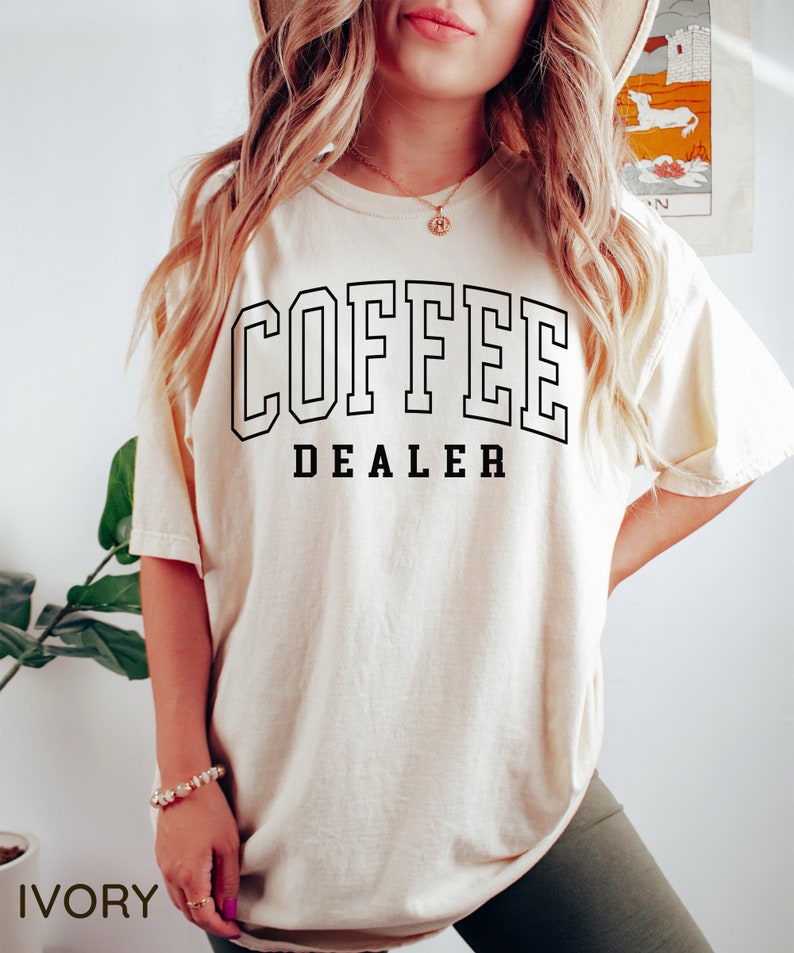 May include: Ivory-colored t-shirt with the words "COFFEE DEALER" in black block letters. The text is outlined in black. The shirt is a relaxed fit.