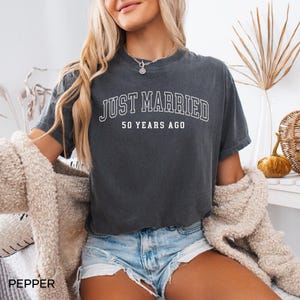 May include: Dark gray t-shirt with the text "JUST MARRIED 50 YEARS AGO" in white. The shirt is a short-sleeved, crew neck style. The word "PEPPER" is printed at the bottom left of the image.