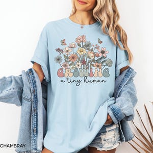 May include: Light blue t-shirt with a floral design and the text "GROWING a tiny human" in pastel colors. The shirt features a bouquet of flowers in shades of pink, yellow, and peach, with butterflies. The model is wearing a denim jacket.