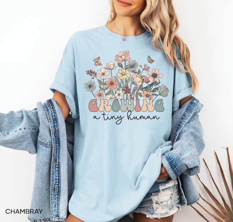 May include: Light blue t-shirt with a floral design and the text "GROWING a tiny human" in pastel colors. The shirt features a bouquet of flowers in shades of pink, yellow, and peach, with butterflies. The model is wearing a denim jacket.