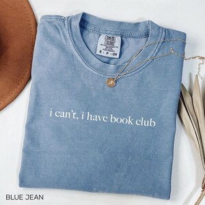 May include: A light blue t-shirt with the text "i can't, i have book club" printed in white. The shirt is made of a soft, comfortable material.