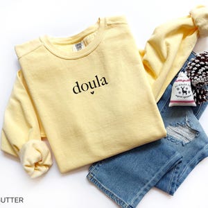 May include: A pale yellow long-sleeved sweatshirt with the word "doula" printed in black. The sweatshirt is paired with blue jeans and a small decorative bag. The word "BUTTER" is printed at the bottom of the image.