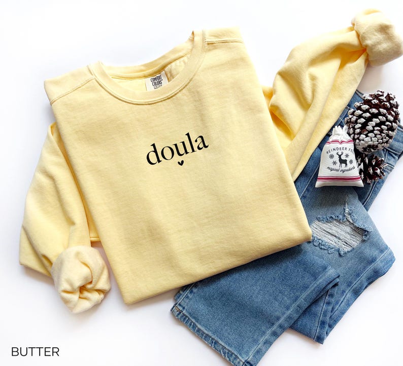 May include: A pale yellow long-sleeved sweatshirt with the word "doula" printed in black. The sweatshirt is paired with blue jeans and a small decorative bag. The word "BUTTER" is printed at the bottom of the image.