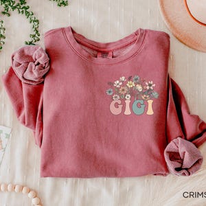 以下が含まれることがあります： 花柄のデザインとカラフルな文字で「GIGI」と書かれた、マルーンのトレーナーです。