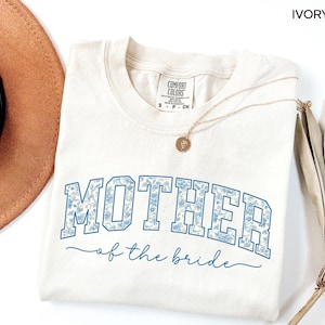 May include: Ivory-colored t-shirt with the words "MOTHER" and "of the bride" in blue floral print. The shirt has a crew neck and short sleeves. A gold necklace with a pendant is draped across the shirt.