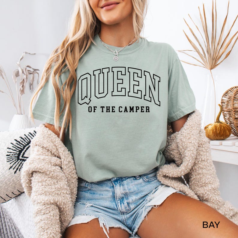May include: A light green t-shirt with the text "QUEEN OF THE CAMPER" in black lettering. The shirt is worn by a person wearing denim shorts.