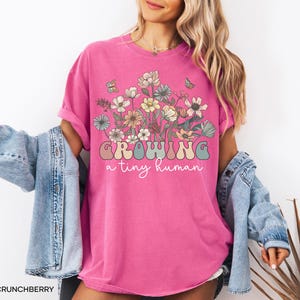 May include: A bright pink t-shirt with the words "GROWING a tiny human" in a retro font, and a floral graphic. The shirt is a crew neck and has rolled-up sleeves. The model is wearing a denim jacket.
