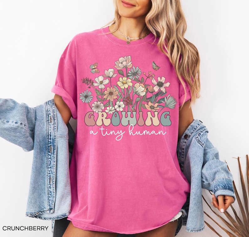 May include: A bright pink t-shirt with the words "GROWING a tiny human" in a retro font, and a floral graphic. The shirt is a crew neck and has rolled-up sleeves. The model is wearing a denim jacket.
