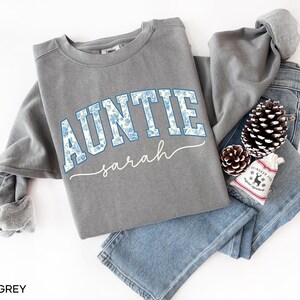 May include: A grey sweatshirt with the word "AUNTIE" in blue floral print lettering, with the name "sarah" in a cursive font underneath. The sweatshirt is folded on top of a pair of blue jeans, with a pinecone and a small decorative bag.