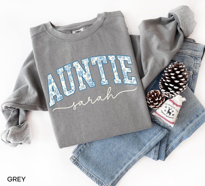 May include: A grey sweatshirt with the word "AUNTIE" in blue floral print lettering, with the name "sarah" in a cursive font underneath. The sweatshirt is folded on top of a pair of blue jeans, with a pinecone and a small decorative bag.