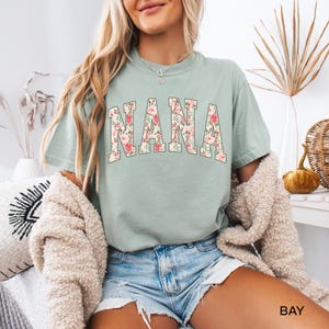 May include: A light green t-shirt with the word "NANA" in large, floral-patterned letters. The shirt is a relaxed fit and has short sleeves. The text "BAY" is visible in the bottom right corner.