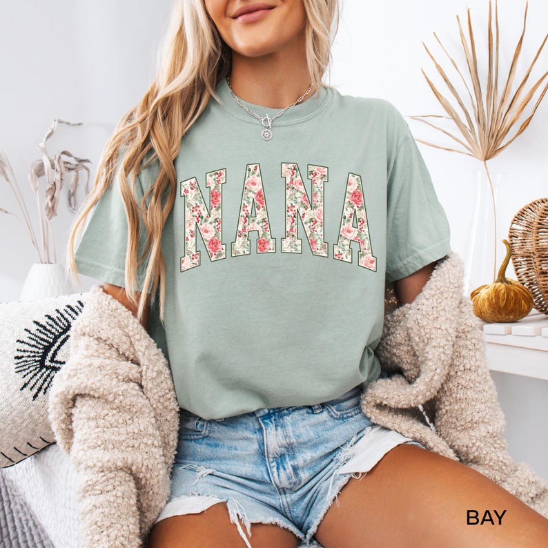 May include: A light green t-shirt with the word "NANA" in large, floral-patterned letters. The shirt is a relaxed fit and has short sleeves. The text "BAY" is visible in the bottom right corner.