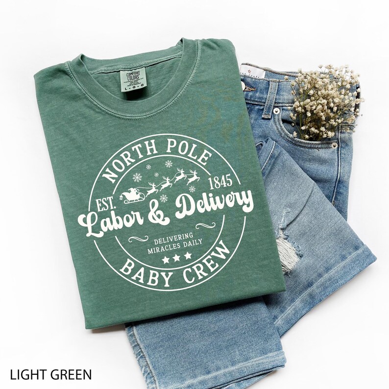 May include: A light green t-shirt with the text "NORTH POLE Labor & Delivery BABY CREW" in a circular design. The design includes snowflakes, reindeer, and a sleigh. The shirt is paired with blue denim shorts and a small bouquet of white flowers.