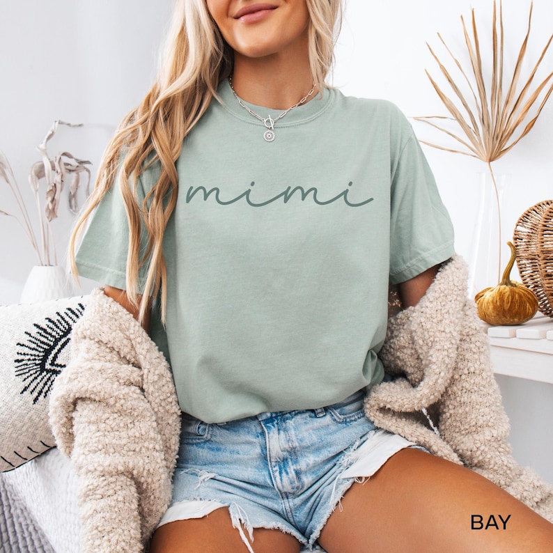 May include: A light green t-shirt with the word "mimi" written in cursive on the front. The shirt is being worn by a person with long blonde hair and is paired with denim shorts.