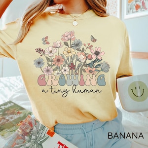 May include: A light yellow t-shirt with a floral design and the text "GROWING a tiny human". The graphic features pastel-colored flowers and butterflies. The word "BANANA" is printed in the bottom right corner.