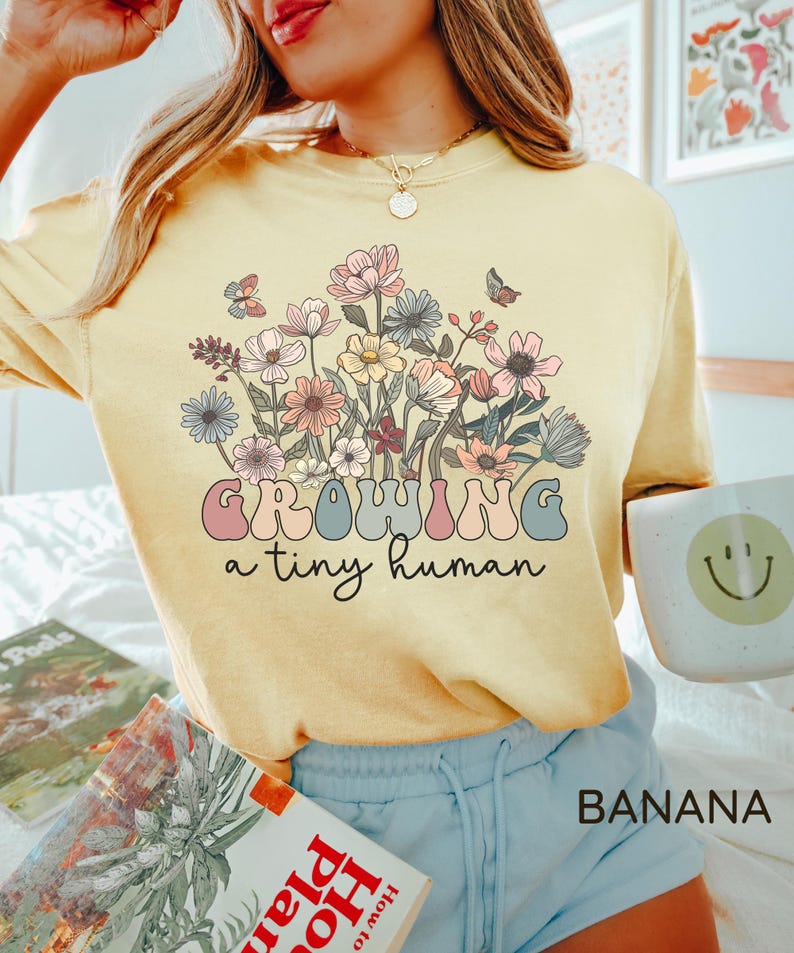 May include: A light yellow t-shirt with a floral design and the text "GROWING a tiny human". The graphic features pastel-colored flowers and butterflies. The word "BANANA" is printed in the bottom right corner.