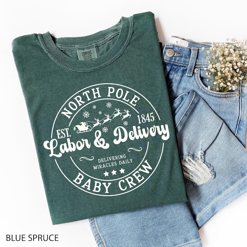 May include: A blue spruce t-shirt with a white circular graphic that reads "North Pole Labor & Delivery Baby Crew" with reindeer and a sleigh. The shirt is paired with denim jeans and a small bouquet of white flowers.