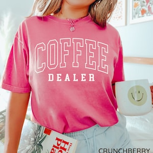 May include: A bright pink t-shirt with the words "COFFEE DEALER" in white block letters. The person is holding a white mug with a yellow smiley face and a book titled "How to Grow House Plants". The word "CRUNCHBERRY" is at the bottom.