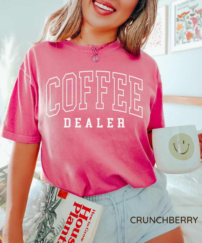 May include: A bright pink t-shirt with the words "COFFEE DEALER" in white block letters. The person is holding a white mug with a yellow smiley face and a book titled "How to Grow House Plants". The word "CRUNCHBERRY" is at the bottom.